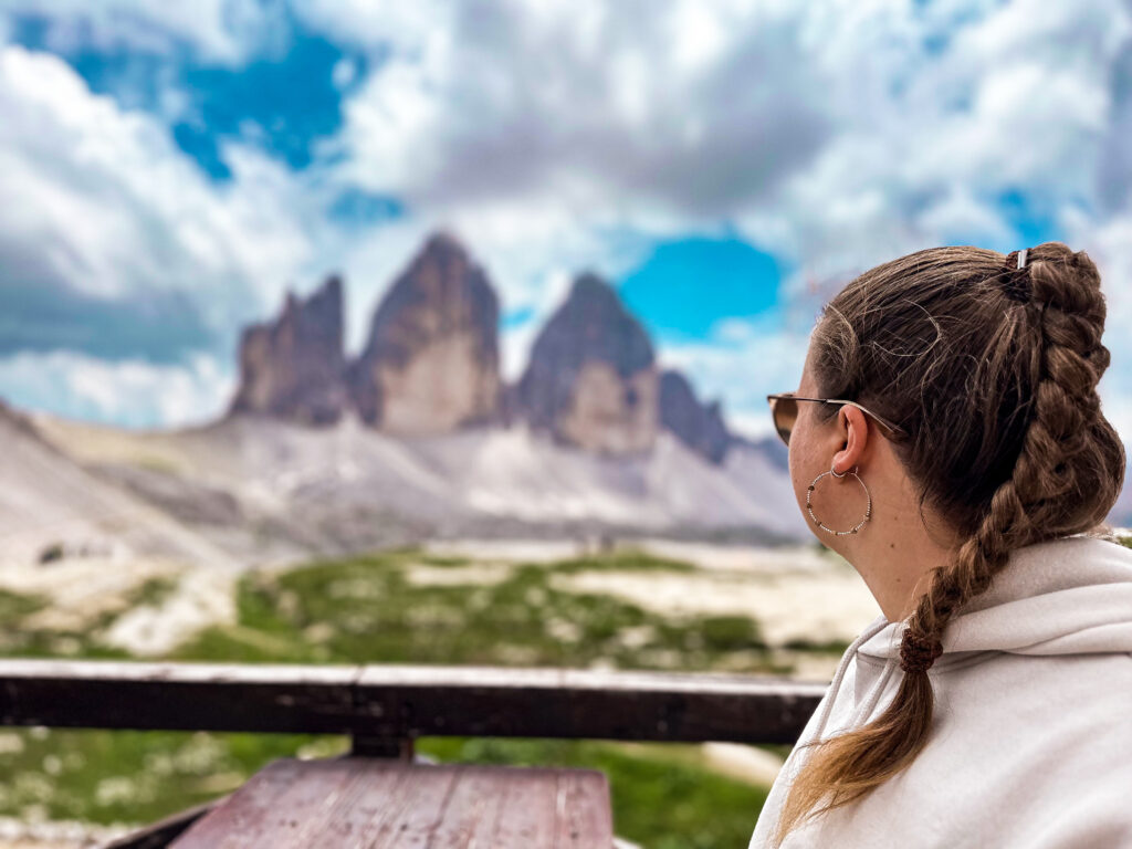Een vrouw met een vlecht kijkt uit over het iconische berglandschap van de Drei Zinnen in de Dolomieten, Italië. De lucht is helder met wolken, en de majestueuze bergtoppen steken scherp af tegen de blauwe lucht.